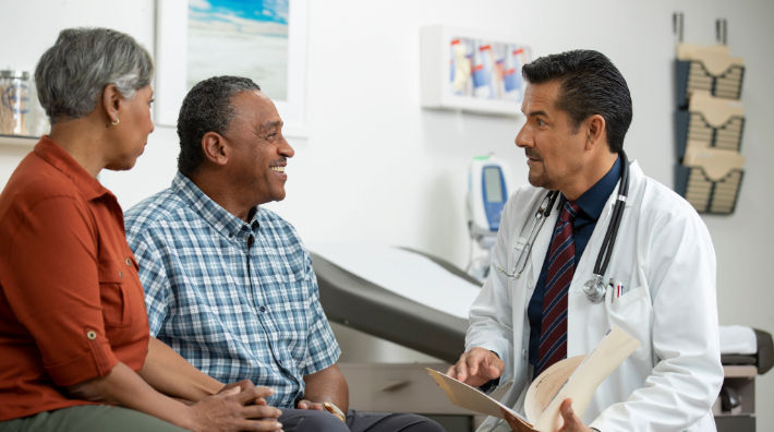 A gray-haired woman holds hands with a man as a male doctor wearing a lab coat talks to them in an exam room at a doctor’s office. 